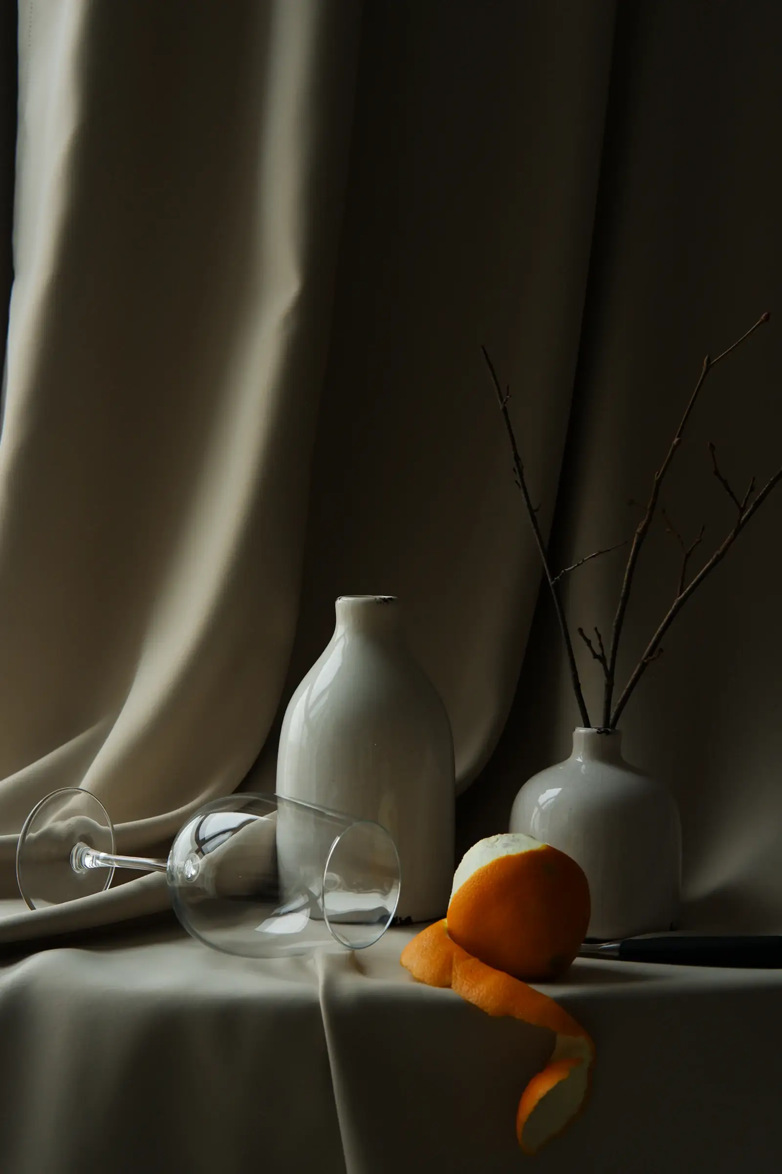 Still life of white ceramic vases, a peeled orange and a tipped wine glass on draped linen in soft shadow